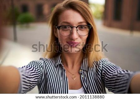 Close up portrait of attractive hipster girl in optical spectacles making selfie photos during leisure time at urban setting, beautiful woman in glasses photographing herself during free day outdoors