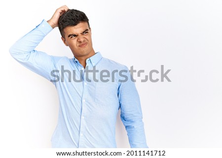 Young hispanic man wearing business shirt standing over isolated background confuse and wondering about question. uncertain with doubt, thinking with hand on head. pensive concept. 