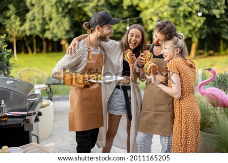Young people enjoy yummy burgers, made on a grill at picnic, standing together and having fun. Friends cooking at backyard outdoors. American lifestyle