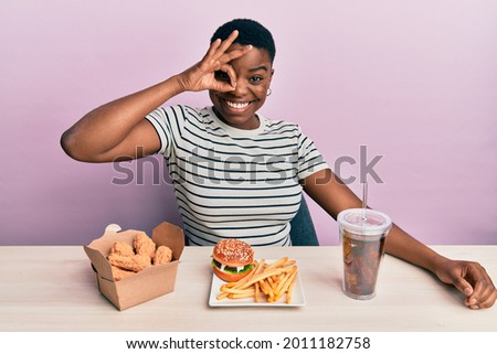 Young african american woman eating a tasty classic burger with fries and soda smiling happy doing ok sign with hand on eye looking through fingers 