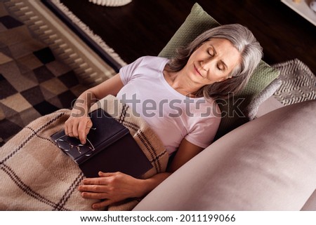 Photo of cute dreamy retired woman dressed white t-shirt lying couch sleeping reading book smiling indoors flat home house
