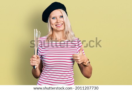 Young caucasian woman holding paintbrushes smiling happy and positive, thumb up doing excellent and approval sign 