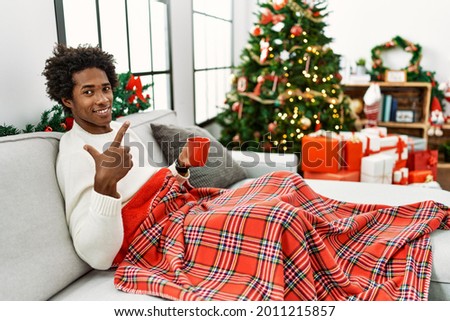 Young african american man sitting on the sofa drinking coffee by christmas tree cheerful with a smile on face pointing with hand and finger up to the side with happy and natural expression 
