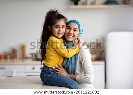 Portrait Of Happy Muslim Mother In Hijab And Little Daughter Posing In Kitchen Interior, Cute Small Girl Hugging Her Islamic Arab Mom And Smiling At Camera, Mommy And Child Having Fun At Home