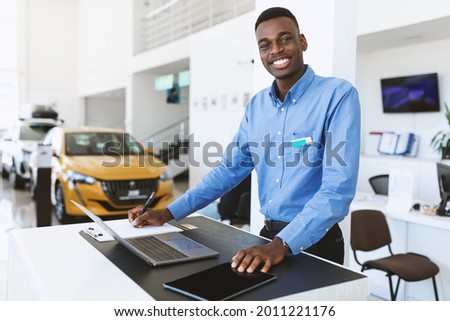 Portrait of happy car salesman standing at work desk, smiling at camera, using laptop and touch pad in auto dealership. Positive black automobile manager posing at workplace in showroom store Royalty-Free Stock Photo #2011221176