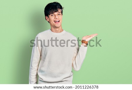 Handsome hipster young man wearing casual winter sweater smiling cheerful presenting and pointing with palm of hand looking at the camera. 