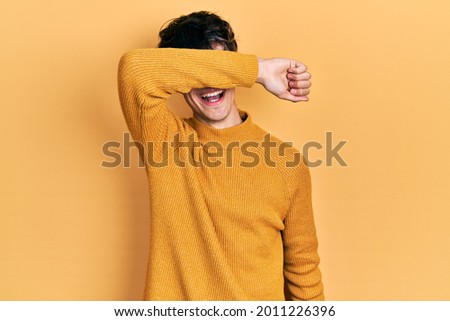 Handsome hipster young man wearing casual yellow sweater covering eyes with arm smiling cheerful and funny. blind concept. 