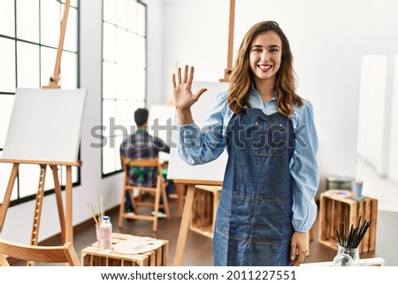 Young artist woman at art studio showing and pointing up with fingers number five while smiling confident and happy. 