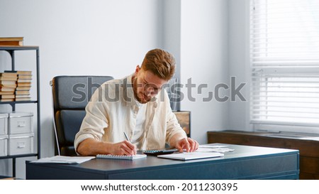 Bearded accountant sums up unexpected expenses from checks on calculator and writes results in paper notebook sitting in office Royalty-Free Stock Photo #2011230395
