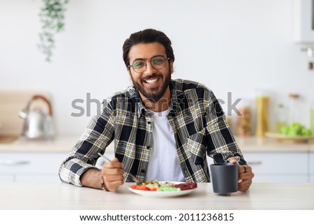 Cheerful middle-eastern young bearded man in casual outfit and stylish glasses enjoying healthy lunch at home, sitting at kitchen table, eating vegetable salad and drinking tea, healthy deit concept Royalty-Free Stock Photo #2011236815