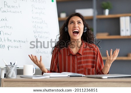 Fatigued From Work. Tired young teacher in headset screaming out loud, gesturing and looking up, having video lesson with students, tutoring English language online, sitting at desk and yelling Royalty-Free Stock Photo #2011236851