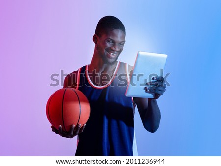Millennial black basketball fan watching game match online, cheering for favorite player or team on tablet pc in neon light. Young African American guy with ball making bet on sports