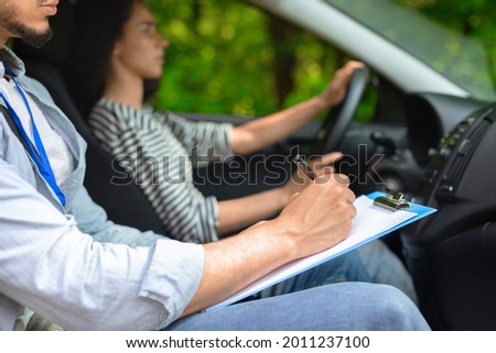Cropped of male instructor examinating female student, taking notes at test chart while sitting by young woman driving automobile, side view, copy space, closeup. Driving school concept