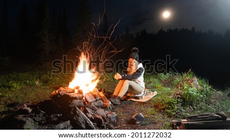 A girl is sitting by a fire at night in the forest. Warms your hands