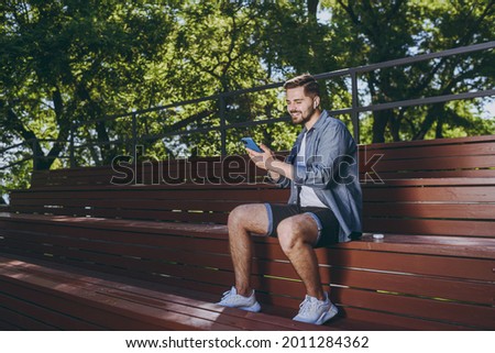 Side view smiling young man in blue shirt shorts earphones sit on bench listen to music using mobile cell phone chat online rest relax in spring green city park outdoor on nature Urban leisure concept