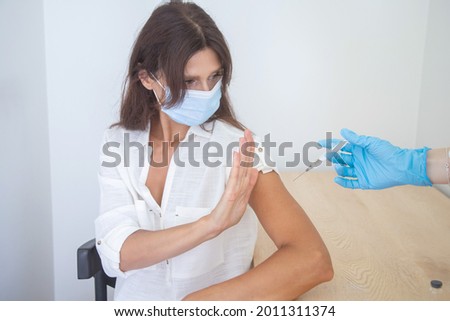 A woman shows with a sign a protest against vaccinations of coronavirus, covid-19, a doctor in blue latex gloves holds a syringe and is ready to inject a patient in the shoulder.
