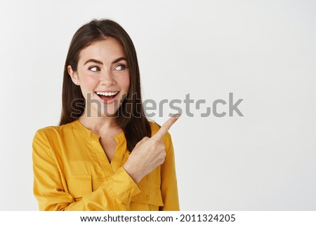 Close-up of lady smiling, pointing and looking right with surprised face, standing on white background