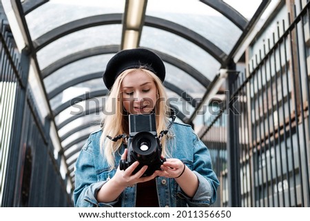 Young woman taking pictures with a medium format film camera. She is wearing a denim jacket and a black beret. Looking to camera.