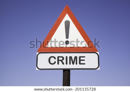 White road warning triangle with black  exclamation point and red frame on  a wooden mast in front of a blue sky. A second rectangular sign warns in english about crime