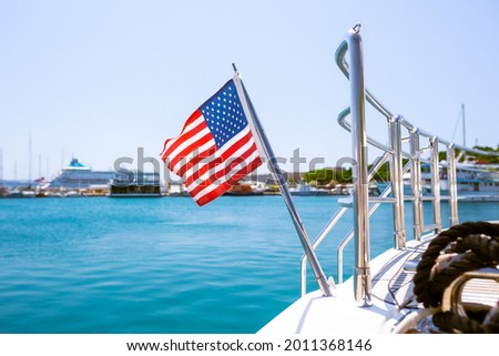 The flag of the United States flutters in the wind on a stainless steel flagpole at the stern of a motor yacht. Marina in the port city in beautiful summer sunny weather. Royalty-Free Stock Photo #2011368146
