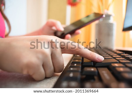 Woman a mobile phone in hand is pressing a key on the computer keyboard by her index finger close up.