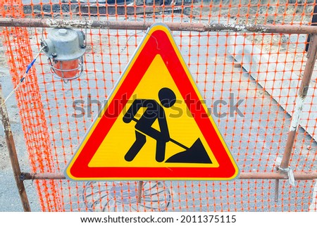 Traffic sign hangs on the fence. Road works are underway. The sign depicts a man with a shovel

