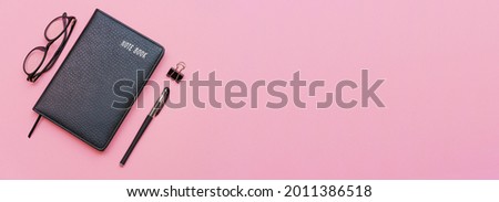 Black notebook with modern glasses and black pen isolated on pink background.