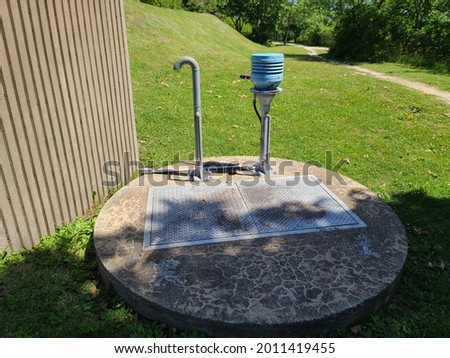 The top of a sewer lift located in Nova Scotia. There are two pipes. One is curved and looks like a cane. The other is capped with a light blue top. The cover is cement and round with a door hatch. Royalty-Free Stock Photo #2011419455