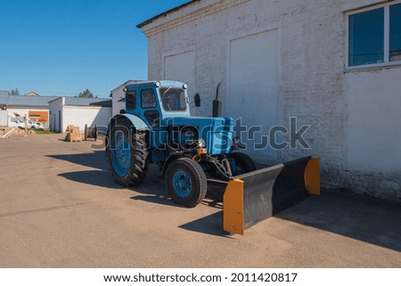 An old blue tractor stands on the asphalt near a white wall. Warehouses are in the background. Sunny weather, bright blue sky.