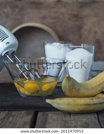 Stage of cooking a banana biscuit. Beat eggs with a mixer in a glass bowl. Ingredients for making biscuit on a brown ancient wooden background. Pastry background
