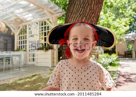 Adorable girl in pirate hat looking at camera and laughing while having fun in courtyard in summer