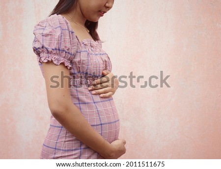 Beautiful pregnant Asian woman touching her belly with hands on wall background.