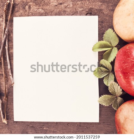 Top view photo of autumn composition on wooden background with white blanc sheet of paper, branches, green strawberry leaves, yellow and red apples. Warm atmosphere.