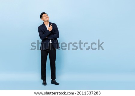 Full length portrait of young handsome southeast Asian businessman looking up and pointing to copy space on light blue studio background Royalty-Free Stock Photo #2011537283