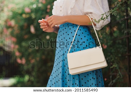 Summer street fashion details: close up of  classic white faux leather bag, small handbag in elegant outfit. Woman wearing trendy polka dot blue midi skirt,  posing in street. Copy, empty  space Royalty-Free Stock Photo #2011585490
