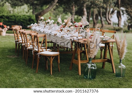 A large, long, decorated, wooden table and chairs, covered with a white tablecloth with dishes, flowers, candles, stands on the green grass in the park, in the forest in nature. Wedding banquet. Royalty-Free Stock Photo #2011627376