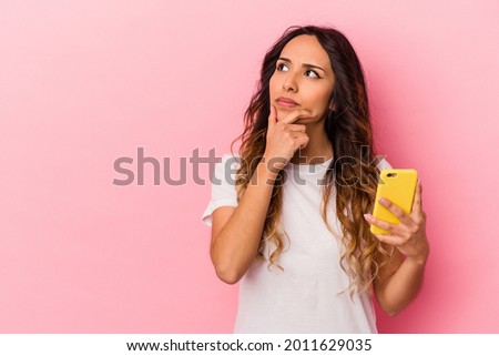 Young mexican woman holding a mobile phone isolated on pink background looking sideways with doubtful and skeptical expression. Royalty-Free Stock Photo #2011629035