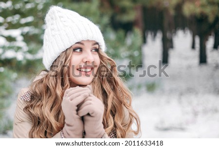 Beautiful girl with white hair stands against the background of the winter forest and smiles. Girl in a white winter hat and mittens does not look at the camera close-up. Concept - new year, Christmas