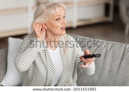 Portrait of confused senior woman with impaired hearing watching TV, trying to listen holding her hand near ear to hear better, sitting on couch in living room. Elderly lady turning volume up Royalty-Free Stock Photo #2011651883