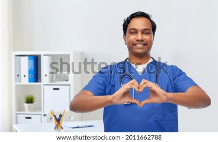 healthcare, profession and medicine concept - happy smiling indian doctor or male nurse in blue uniform with stethoscope showing hand heart over medical office at hospital background