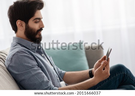 Handsome arab guy sitting with smartphone on sofa, relaxing with cellphone, messaging with friends, checking new app, enjoying mobile communication or browsing social networks, side view