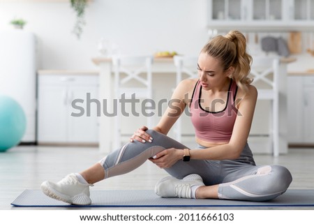 Training injury at home. Woman feeling pain after exercising at home. Desperate young blonde lady in sportswear suffering from knee pain, in minimalist living room, kitchen interior, copy space Royalty-Free Stock Photo #2011666103