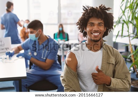 Successful Covid-19 Vaccination Concept. Vaccinated Black Man Gesturing Thumb Up And Showing Arm With Adhesive Bandage After Vaccine Injection, Posing Sitting At Clinic Hospital, Blurred Background Royalty-Free Stock Photo #2011666127