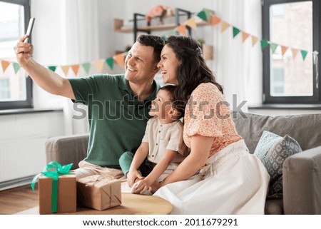 family, holidays and people concept - portrait of happy mother, father and little son with gifts and smartphone taking selfie on birthday at home party