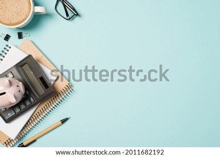 Top view photo of glasses cup of frothy coffee binder clips pen planner and piggy bank on calculator on isolated pastel blue background with copyspace