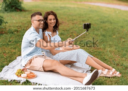 leisure and people concept - happy couple having picnic and taking picture with smartphone on selfie stick at summer park