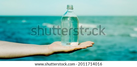 closeup of a young caucasian man holding a glass reusable bottle full of water in the palm of his hand, in front of the ocean, in a panoramic format to use as web banner or header Royalty-Free Stock Photo #2011714016