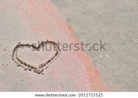The heart is painted on the white and pink sand on the seashore by the water. 