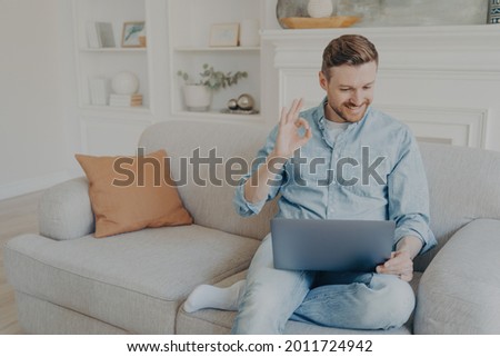 Handsome caucasian male freelancer smiling and showing sign OK with fingers while having video conference on notebook, young man in casual clothes using wireless laptop for online meeting from home