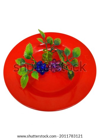 Toy grapes on a red plate isolated white. The plate is made of plastic and is red. On the plate are toy grapes. This plate and toy grapes were photographed from above on a white background.